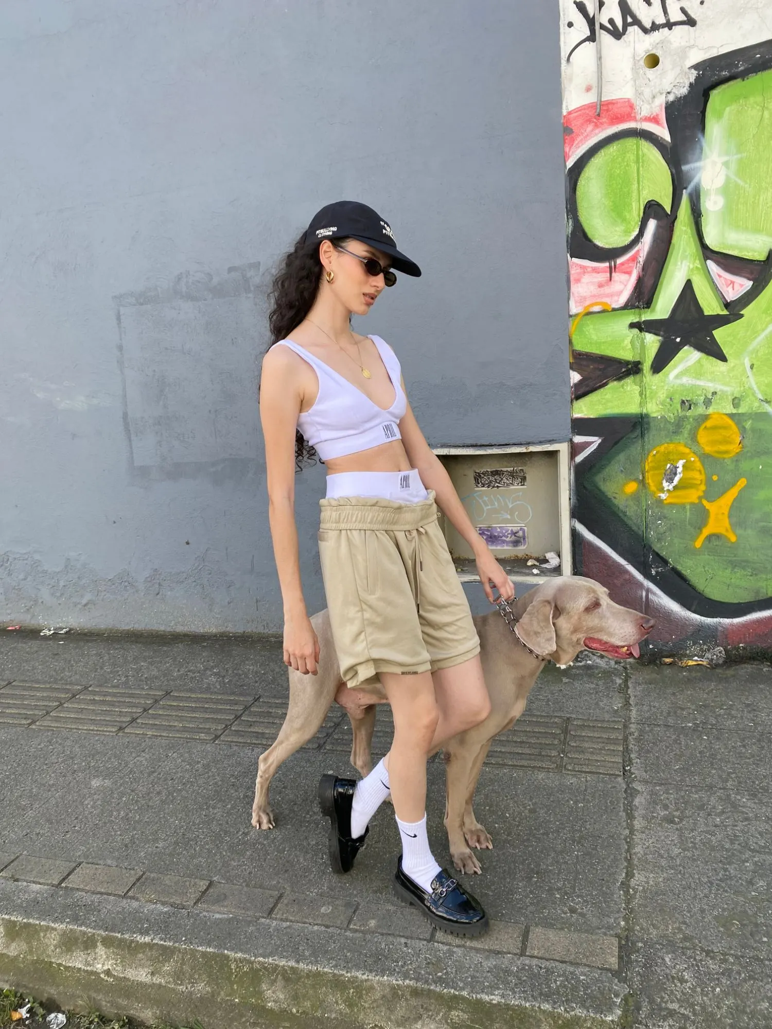 Mujer con gorra negra, gafas pequeñas, top blanco y bermuda beige, paseando un perro weimaraner frente a un grafiti verde.