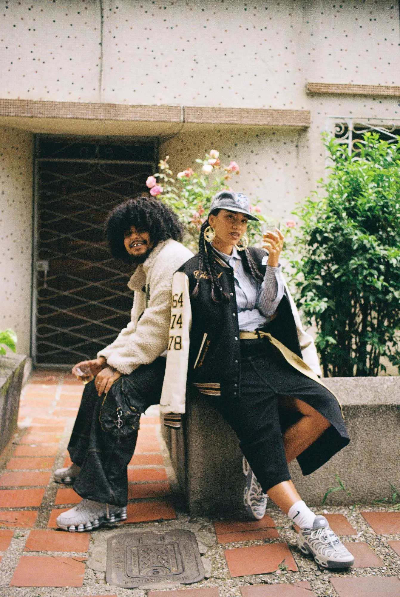 Pareja sentada en el muro de un patio con rosas: él con chaqueta de borrego y jean negro, ella con la varsity, falda negra y tenis plateados.