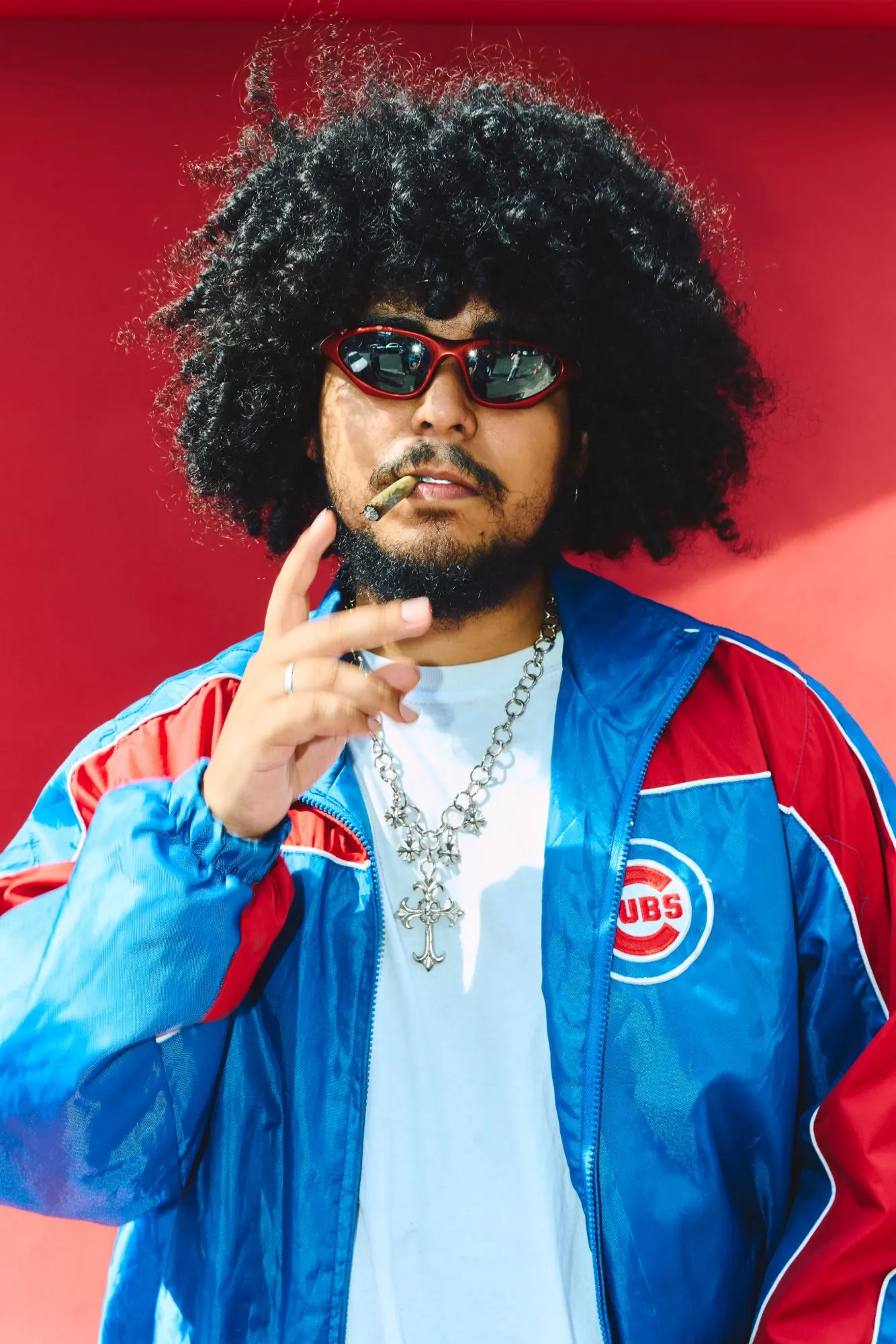 Hombre de pelo afro y gafas rojas, con rompevientos de béisbol azul y rojo y cadena con cruz, contra una pared roja.