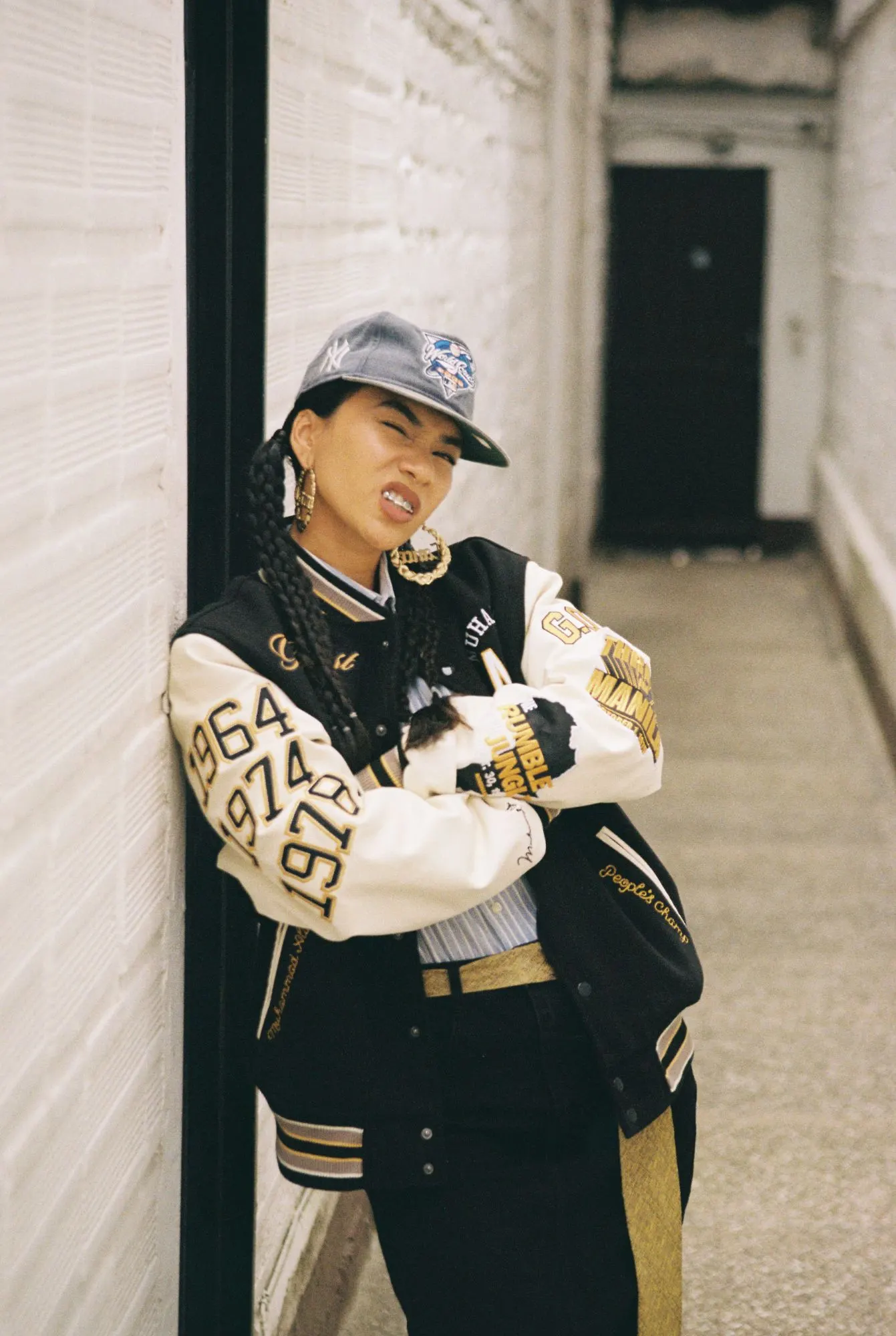 Mujer con gorra gris, trenzas largas, argollas doradas y chaqueta varsity negra y crema, recostada contra una pared blanca de ladrillo.