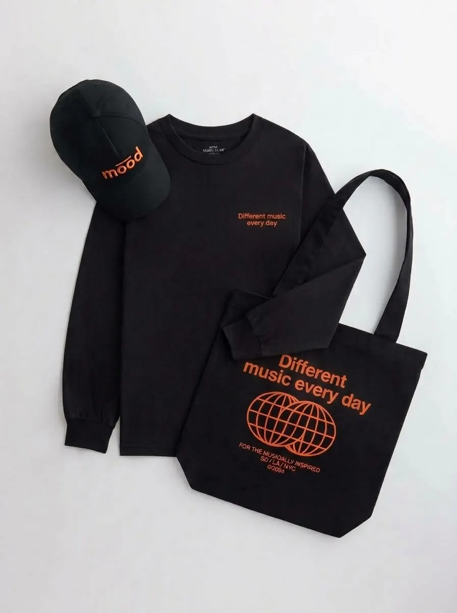 Manga larga negra, gorra negra y tote negra sobre fondo blanco; la gorra dice mood y la manga larga y la tote dicen «Different music every day» en naranja.